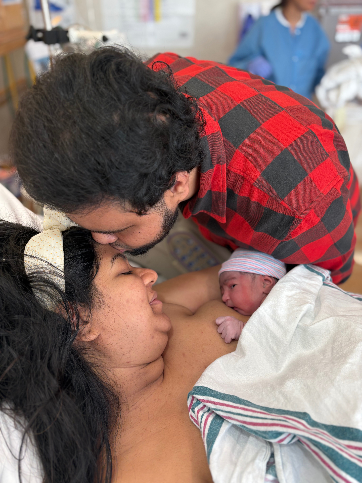 A father kisses the mother's forehead as their newborn rests skin-to-skin